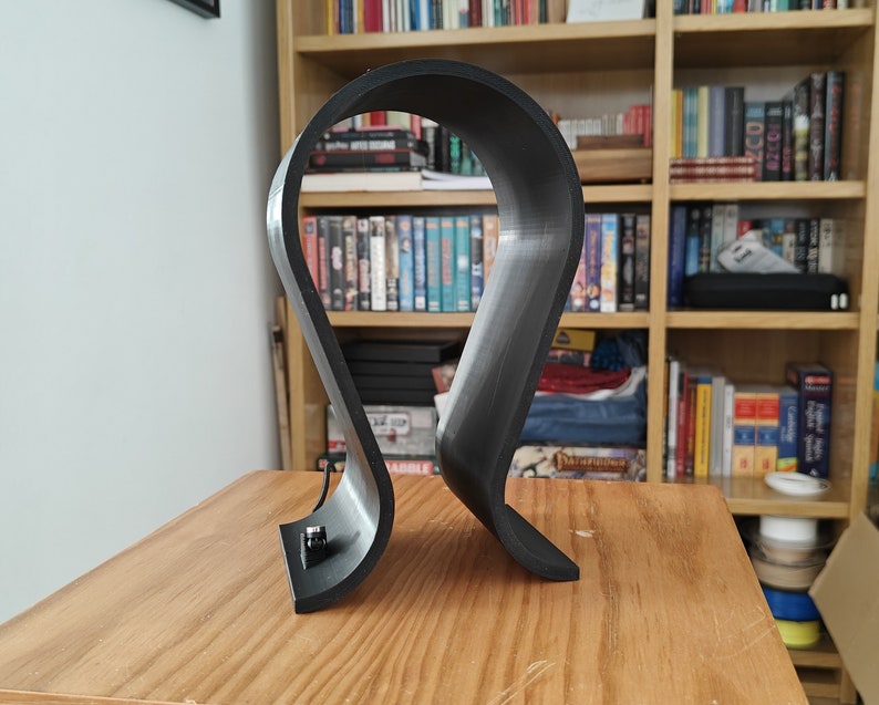 Op de afbeelding: Een zwarte plastic hoofdtelefoonstandaard met een gebogen ontwerp. De standaard staat op een houten oppervlak.