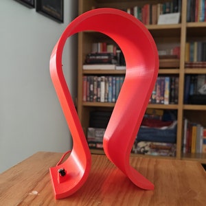 Op de afbeelding: Een felrode 3D-geprinte koptelefoonstandaard. De standaard is gevormd als een grote S en heeft een klein gaatje aan de onderkant voor een oplaadkabel.