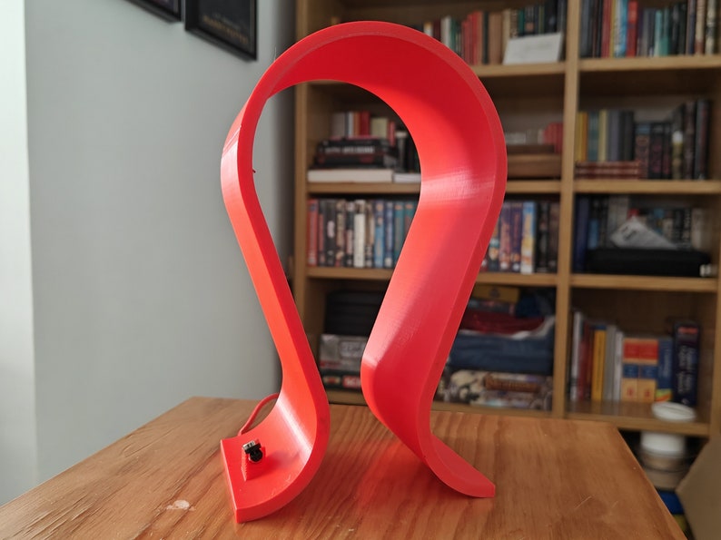 Op de afbeelding: Een felrode 3D-geprinte koptelefoonstandaard. De standaard is gevormd als een grote S en heeft een klein gaatje aan de onderkant voor een oplaadkabel.