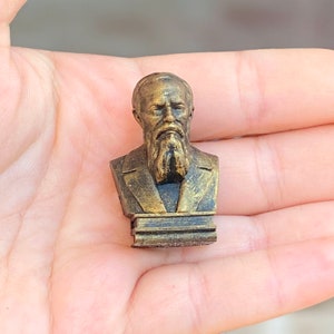 May include: A gold-colored bust of a man with a beard, on a small, rectangular base. The bust is made of a smooth, shiny material.