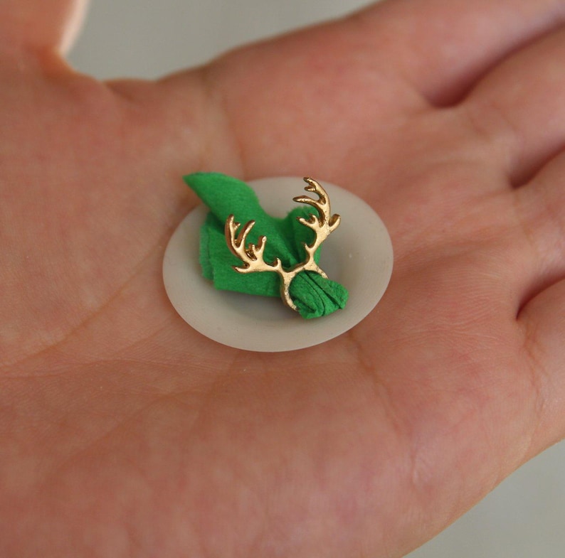 Puede incluir: Un anillo de servilleta de reno dorado que sostiene una servilleta verde en un plato blanco.
