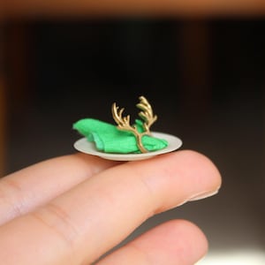 Puede incluir: Un anillo de servilleta de reno dorado en miniatura en un peque&ntilde;o plato blanco con una servilleta verde.
