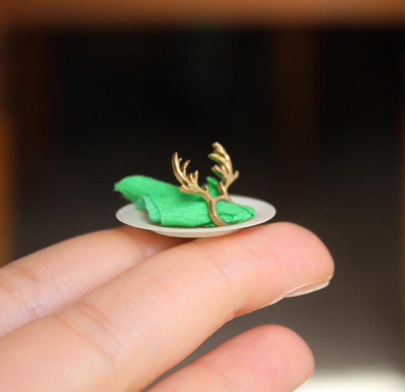 Puede incluir: Un anillo de servilleta de reno dorado en miniatura en un peque&ntilde;o plato blanco con una servilleta verde.
