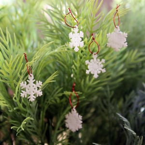 May include: White snowflake ornaments with red hanging loops are displayed on a green, leafy background. The ornaments are in various sizes and shapes, adding a festive touch. The image captures a close-up view of the holiday decorations.