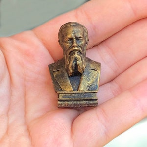 May include: A gold-colored bust of a man with a beard, on a small base. The bust is made of a 3D printed material.