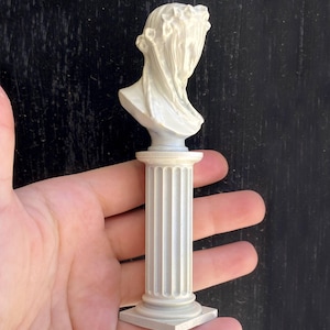 May include: White plaster bust of a woman with a draped cloth over her head, on a white pedestal with fluted columns.