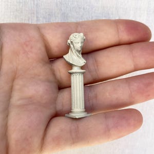 May include: A white plaster bust of a person on a white pedestal. The bust is of a person with their head tilted slightly to the side. The pedestal has a fluted column and a square base.