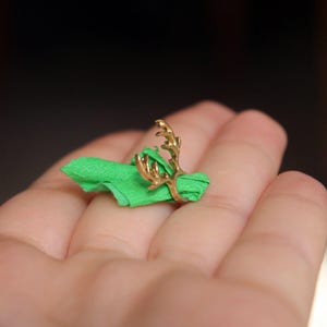 Puede incluir: Un anillo de oro con un dise&ntilde;o de asta de ciervo. El anillo est&aacute; en un dedo que lleva una servilleta de tela verde.