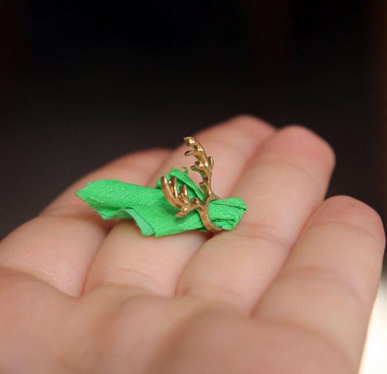 Puede incluir: Un anillo de oro con un dise&ntilde;o de asta de ciervo. El anillo est&aacute; en un dedo que lleva una servilleta de tela verde.