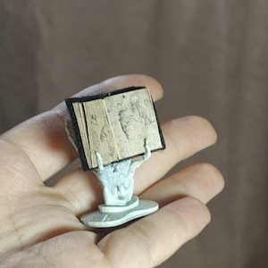 May include: A miniature open book with detailed illustrations, held aloft by a skeletal hand sculpture. The book has a black cover and aged-looking pages. The sculpture is light gray and rests on a small, round base.