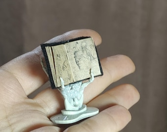 May include: A miniature open book with detailed illustrations, held aloft by a skeletal hand sculpture. The book has a black cover and aged-looking pages. The sculpture is light gray and rests on a small, round base.