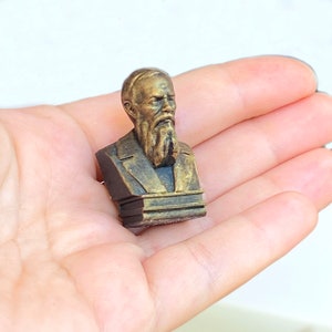 May include: A small, gold-colored bust of a man with a beard, on a wooden base. The bust is a 3D printed replica of a historical figure.