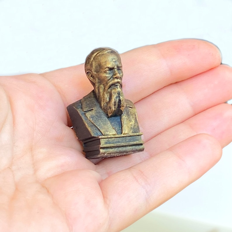May include: A small, gold-colored bust of a man with a beard, on a wooden base. The bust is a 3D printed replica of a historical figure.