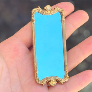 May include: A gold-colored ornate mirror with a blue reflective surface. The mirror is held in a hand.