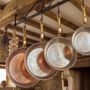 Puede incluir: Sartenes con fondo de cobre y asas de latón cuelgan de un estante de hierro forjado en una cocina rústica. Las sartenes tienen una textura martillada. Cerca cuelgan manojos de hierbas secas y ajo.