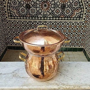 May include: A hammered copper pot with two handles and a lid. The pot is sitting on a white marble surface.