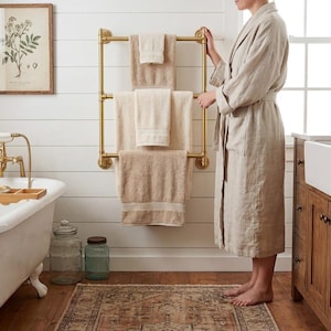 May include: A bathroom interior featuring a roll-top bath, a wooden vanity unit, and a gold towel rail. Beige towels are displayed on the rail. A person in a beige robe stands nearby. A framed botanical print hangs on the wall.