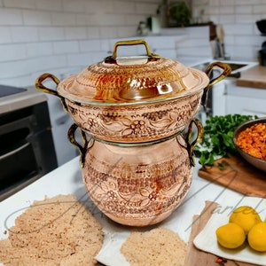 Pode incluir: Uma panela de cozinha de cobre com tampa e desenhos florais intrincados. A panela está empilhada em cima de uma panela de cobre menor com desenhos semelhantes. As panelas estão sobre uma superfície branca com um monte de grãos marrons em primeiro plano.