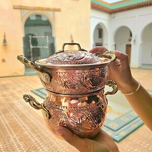Pode incluir: Conjunto de panelas de cobre com desenhos florais e de peixes ornamentados. O conjunto inclui uma panela com tampa e uma panela menor por baixo. As alças e a alça da tampa são douradas. Em um fundo externo desfocado.