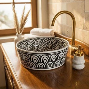 May include: A decorative bathroom sink with a black and white geometric pattern sits on a wooden vanity. A gold faucet and soap dispenser are visible. The sink is a round vessel sink.