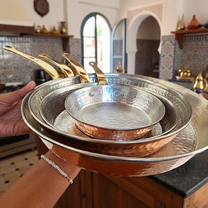 Op de afbeelding: Een set van vier gehamerde koperen pannen met messing handvatten. De pannen zijn in elkaar genest, met de grootste pan op de voorgrond. Gepresenteerd in een keuken.