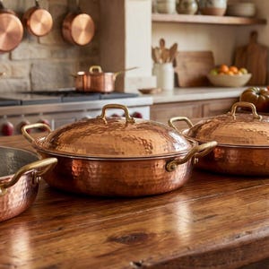 May include: Copper cookware, including pans and lidded pots, arranged on a wooden surface. The kitchen setting features hanging copper pots and a stove in the background. The pans have brass handles.