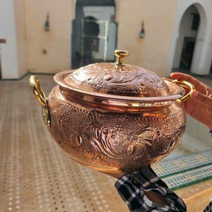 Puede incluir: Una olla de cocina de cobre ornamentada, con tapa y dos asas. La olla presenta intrincados diseños florales y de hojas. La tapa tiene un pequeño asa redonda. La olla es sostenida por una persona que lleva una camisa a cuadros blancos y negros.