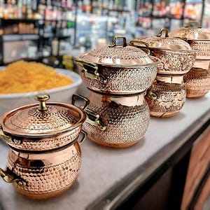 May include: A collection of copper cooking pots with lids, arranged on a countertop. The pots have a hammered texture and brass handles. The background is blurred, suggesting a restaurant or kitchen setting.