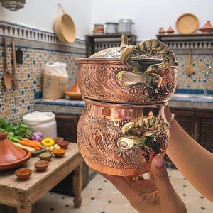 Könnte beinhalten: Verzierte Kochtopf aus Kupfer mit Deckel und dekorativen Griffen. Der Topf hat ein geprägtes Blumenmuster. Das Bild ist in einer Küche mit einem rustikalen Holztisch und einem Tajine-Topf aufgenommen.
