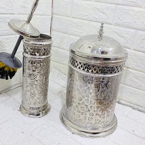 Puede incluir: Dos accesorios de baño plateados. Uno es un portacepillos de inodoro con tapa y cepillo. El otro es un recipiente a juego con tapa. Ambos tienen intrincados patrones en relieve y un diseño decorativo.