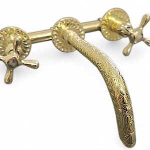 May include: A gold-colored faucet with a curved spout and cross-shaped handles. The faucet features ornate, engraved floral patterns on the spout and around the handles, creating a vintage aesthetic. The fixture is set against a white background.