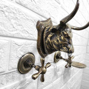 May include: A bronze-toned wall-mounted faucet with a bull head design. The faucet features a detailed bull head with curved horns and decorative elements. The fixture is mounted on a white brick wall, showcasing its antique aesthetic.