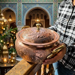 May include: A round copper pot with a matching lid, featuring intricate embossed floral and leaf patterns across the surface. The pot includes two polished gold-tone metal handles on the sides and a small gold-tone knob on the centre of the lid.