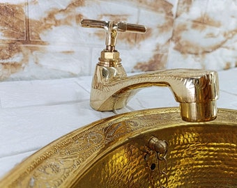 May include: A gold-toned brass sink with an ornate design and a matching faucet. The sink is round and has a hammered texture. The faucet has a simple design with a cross-shaped handle.