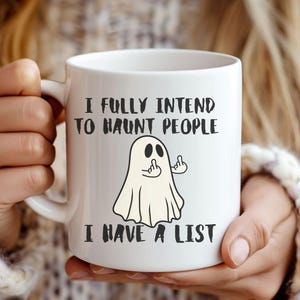 Puede incluir: Taza de cer&aacute;mica blanca con el texto "I FULLY INTEND TO HAUNT PEOPLE I HAVE A LIST" y un fantasma de dibujos animados haciendo un gesto grosero. La taza tiene un asa blanca y la sostiene una persona.