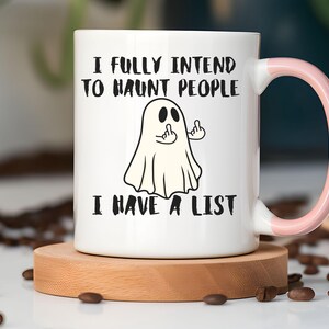 Puede incluir: Taza de cer&aacute;mica blanca con asa rosa, con una ilustraci&oacute;n de un fantasma haciendo una peineta. El texto en la taza dice: "I FULLY INTEND TO HAUNT PEOPLE I HAVE A LIST". La taza est&aacute; sobre un posavasos de madera.