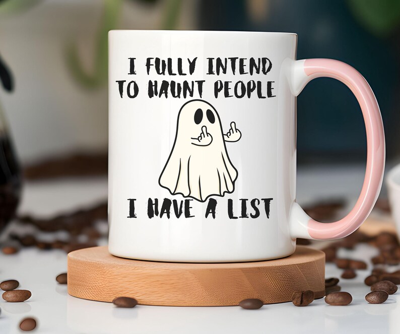 Puede incluir: Taza de cer&aacute;mica blanca con asa rosa, con una ilustraci&oacute;n de un fantasma haciendo una peineta. El texto en la taza dice: "I FULLY INTEND TO HAUNT PEOPLE I HAVE A LIST". La taza est&aacute; sobre un posavasos de madera.