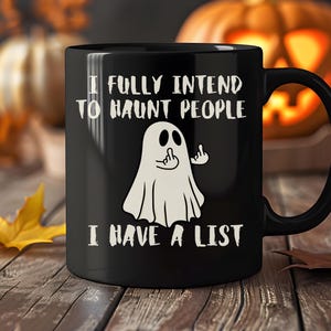 Puede incluir: Taza de cer&aacute;mica negra con el texto "I FULLY INTEND TO HAUNT PEOPLE I HAVE A LIST" y un fantasma de dibujos animados haciendo una peineta. La taza tiene un asa negra y est&aacute; sobre un fondo borroso con calabazas y hojas de oto&ntilde;o.