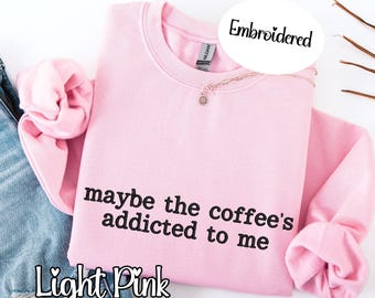 May include: Light pink embroidered sweatshirt with the phrase "maybe the coffee's addicted to me" in black lettering. The sweatshirt is folded, with the sleeves and collar visible. A delicate necklace is draped on the garment.