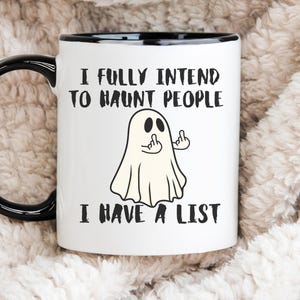 Puede incluir: Taza de cer&aacute;mica blanca con asa y borde negros. La taza presenta una ilustraci&oacute;n de fantasma de dibujos animados con el texto "I FULLY INTEND TO HAUNT PEOPLE I HAVE A LIST."