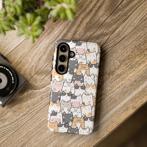 May include: A phone case featuring a repeating pattern of cartoon cats in various colors, including gray, white, orange, and pink. Some cats wear glasses. The case is on a wooden surface.