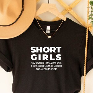 Könnte beinhalten: Schwarzes T-Shirt mit weißem Text, der lautet: "SHORT GIRLS GOD ONLY LETS THINGS GROW UNTIL THEY'RE PERFECT, SOME OF US DIDN'T TAKE AS LONG AS OTHERS".