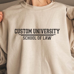 Könnte beinhalten: Ein beiger Pullover mit dem Aufdruck "CUSTOM UNIVERSITY SCHOOL OF LAW" auf der Vorderseite.