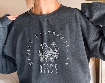 Puede incluir: Una sudadera gris oscura con un diseño gráfico blanco que dice "Easily Distracted By Birds" con una ilustración estilizada de un pájaro.