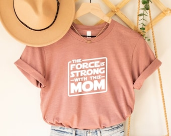 May include: A dusty rose t-shirt with the text "THE FORCE IS STRONG WITH THIS MOM" in white. The shirt is hanging on a wooden hanger, with a tan hat and a gold necklace visible. The shirt is rolled up at the sleeves.