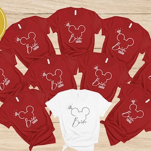 Op de afbeelding: Rode T-shirts met een witte omtrek van Mickey Mouse oren met een diamant erop en de tekst "Bride Squad". Een wit T-shirt met een zwarte omtrek van Mickey Mouse oren met een diamant erop en de tekst "Bride".