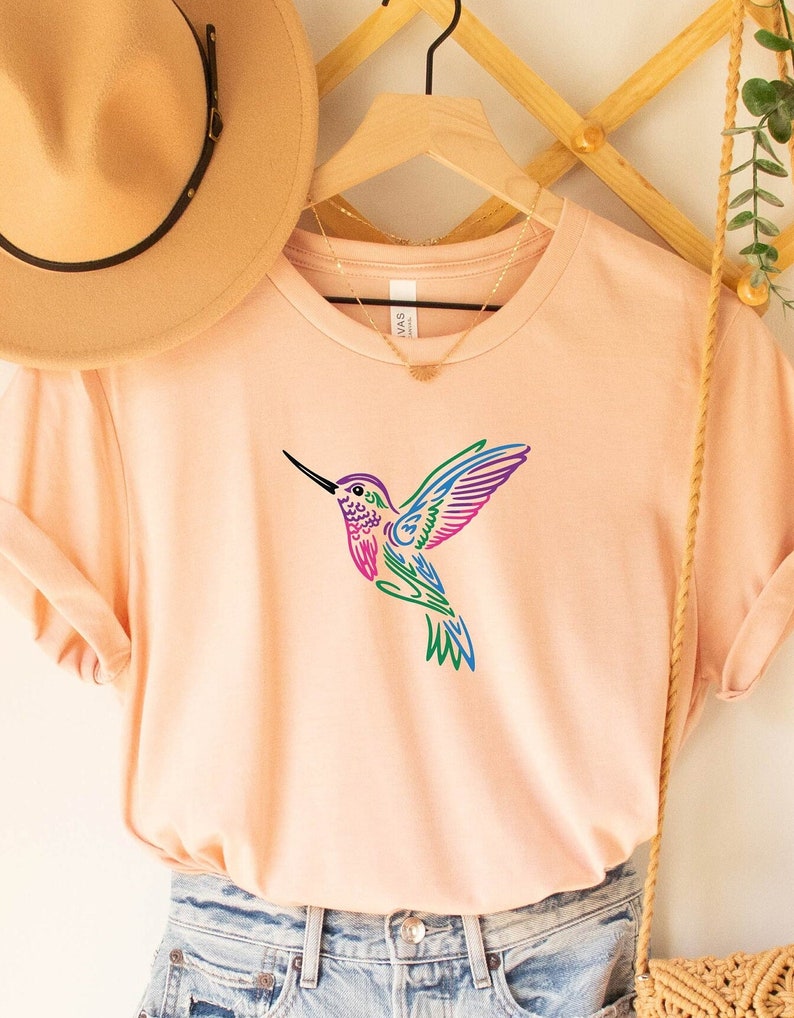 May include: A light pink t-shirt with a colorful hummingbird graphic. The hummingbird is outlined in pink, blue, and green.