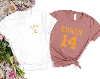 May include: Two white t-shirts with the name "EDWIN" printed on them. One shirt has a hockey stick design and the other has the number "14" printed on it.