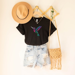 May include: Black t-shirt with a colorful hummingbird graphic. The shirt is hanging on a wooden hanger with a straw hat and a woven bag.