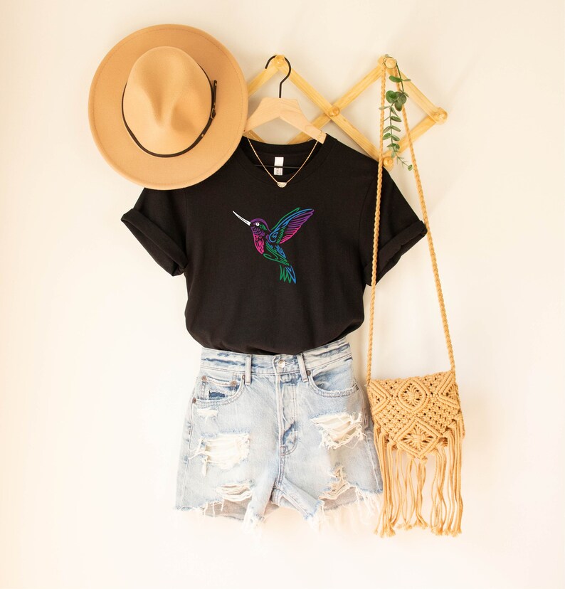 May include: Black t-shirt with a colorful hummingbird graphic. The shirt is hanging on a wooden hanger with a straw hat and a woven bag.
